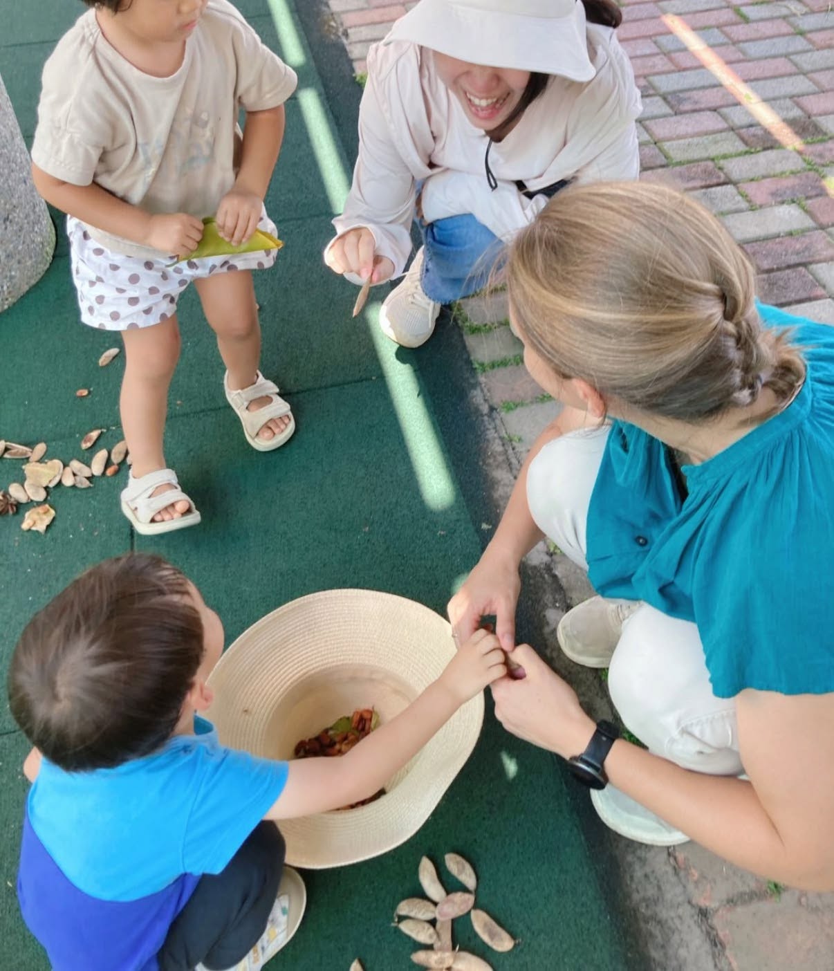 家長蹲下陪孩子一起觀察與收集自然素材，孩子把小小的發現放進帽子裡