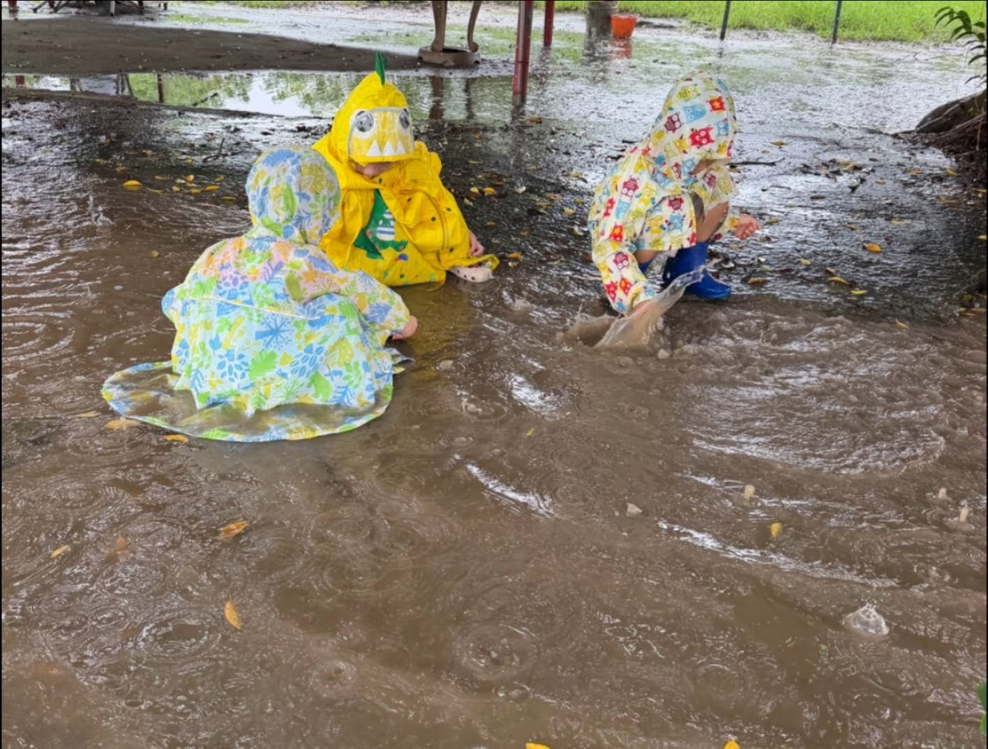 三位穿著彩色雨衣的孩子，在戶外蹲下感受雨水與水窪